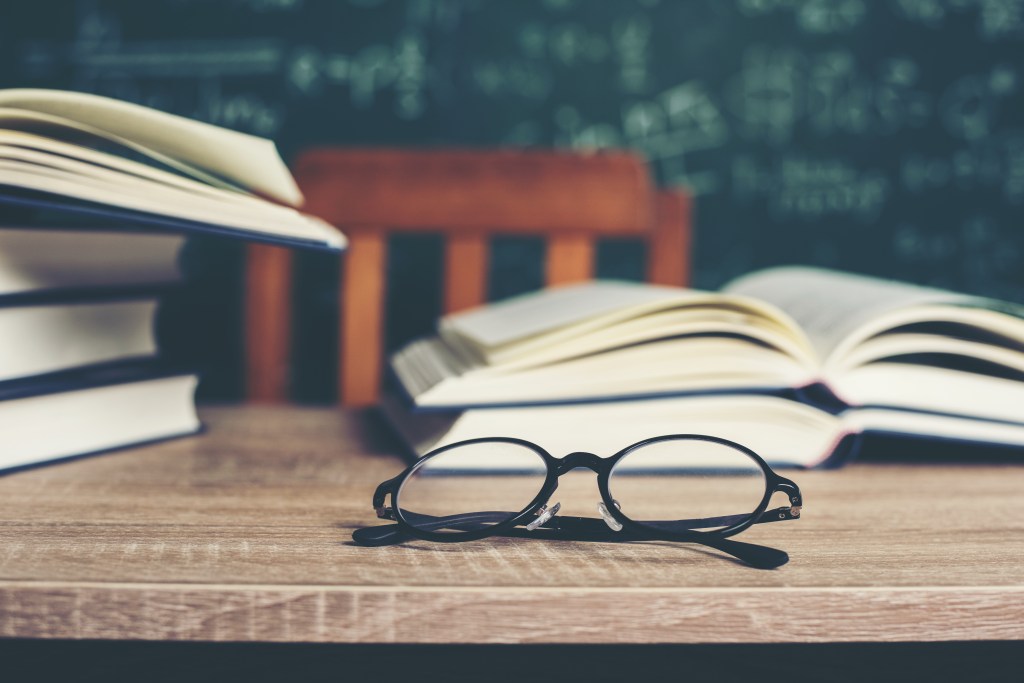 Books and spectacles on desk in front of blackboard