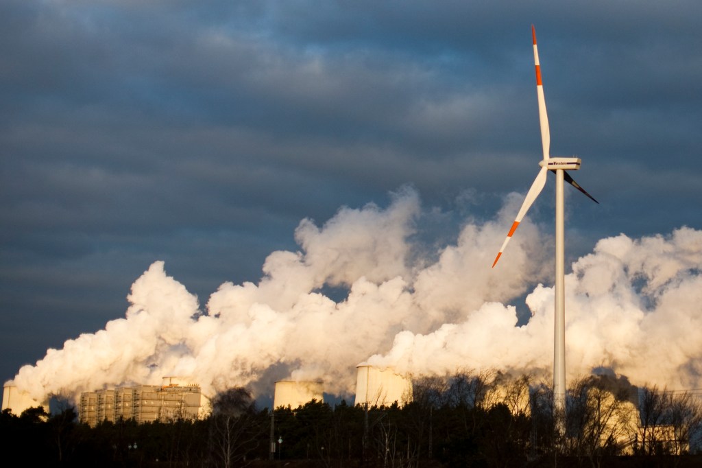 A wind turbine against a backdrop of smoke from factory stacks.