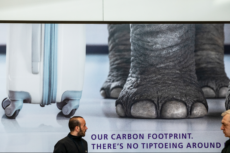 Air passengers walk past a poster advertising Heathrow's commitment to reducing carbon emissions