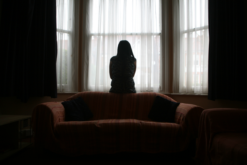 A young woman suffering from domestic violence stands alone in the bay window of her home.