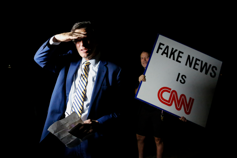 A man holding a sign that reads Fake News is CNN stands next to Michael Williams, an anchor of Channel 5 WPTV (NBC affiliated) as he waits to go live outside of the home of former President Donald Trump at Mar-A-Lago on August 8, 2022 in Palm Beach, Florida. The FBI raided the home to retrieve classified White House documents.