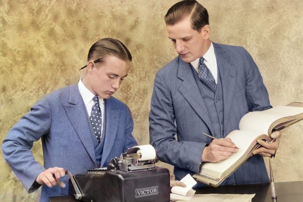 Old-fashioned image of a man and boy bookkeeper using a ledger book.