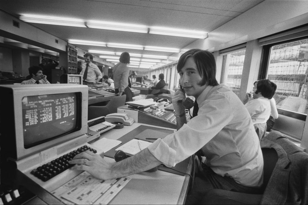 Image of a man in 1970s clothing and hair style near a computer and keyboard, also from that decade.