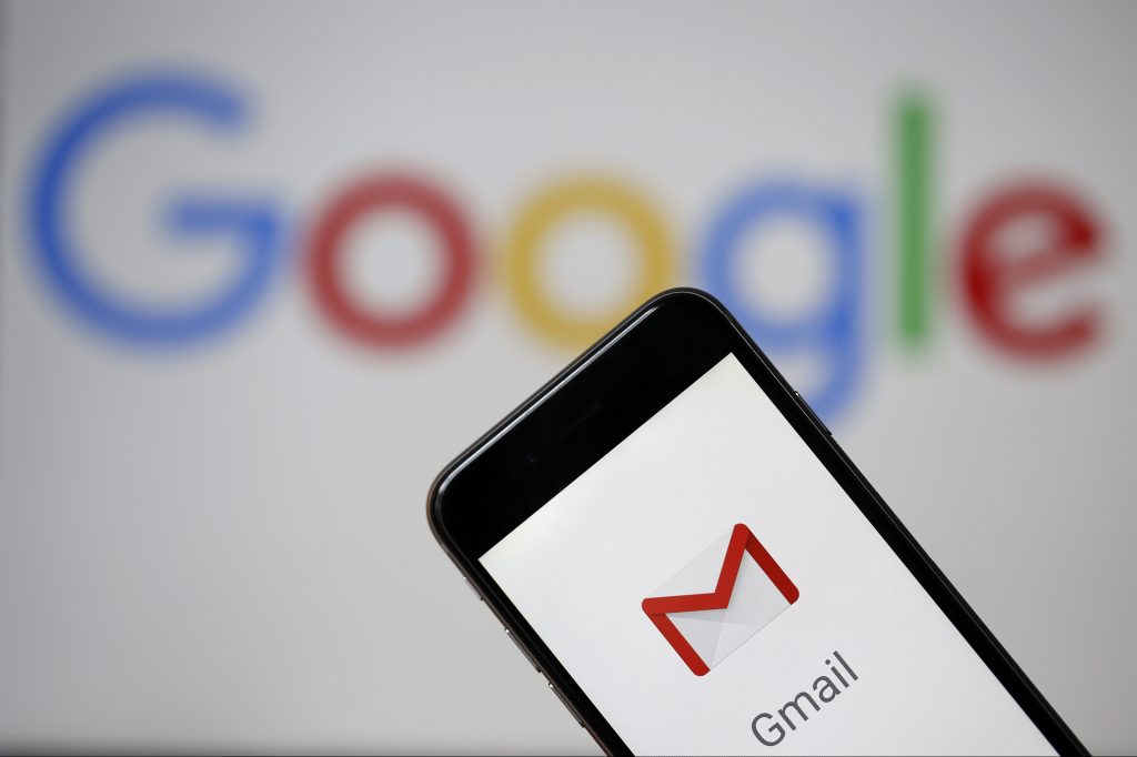 The logo of the Gmail app homepage is seen on the screen of an iPhone in front of a computer screen showing a Google.