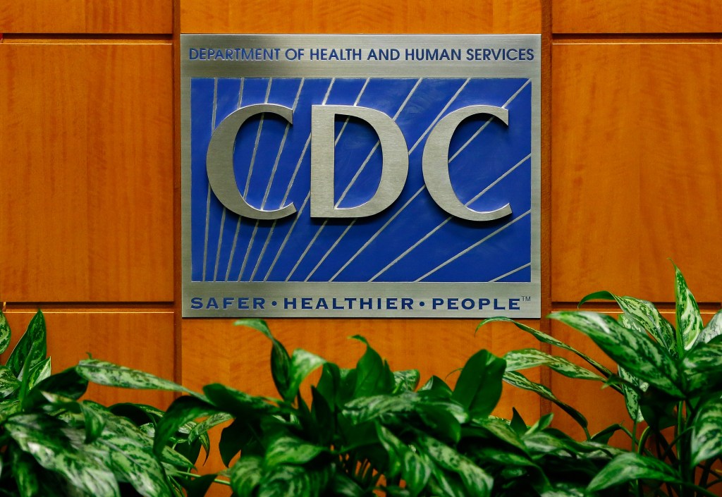 A photo of the Centers for Disease Control and Prevention logo with CDC in gray metal on a blue background mounted on a wood panel wall.