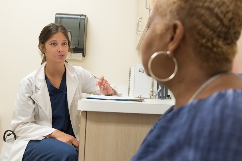 A photo of a seated white, female doctor in a lab coat and blue scrubs speaking with a seated black female patient wearing a blue top in an examination room.