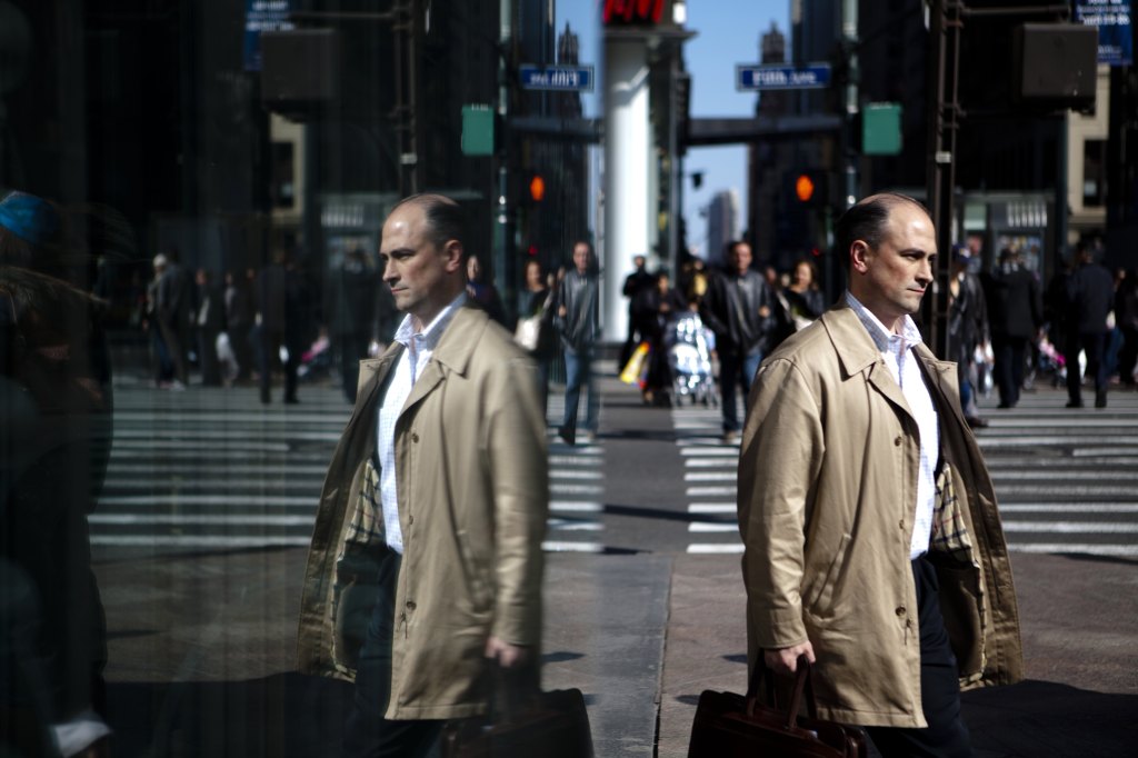 A double image photo showing 2 images of a white man in a rumpled trench coat carrying a briefcase walking in opposite directions on a side walk in New York City.