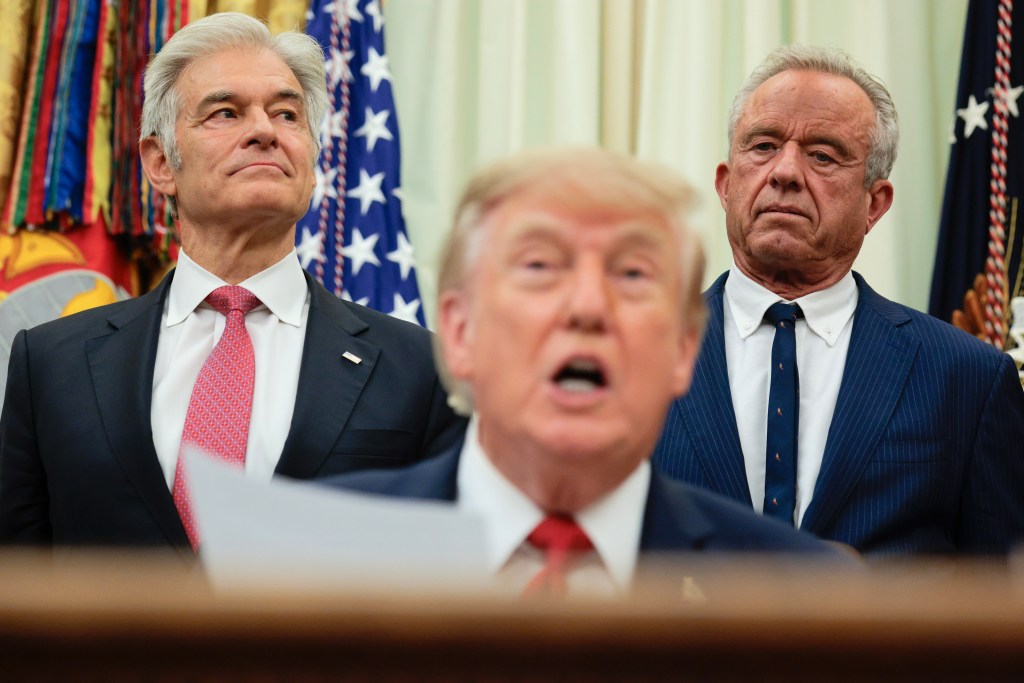 A photo of President Donald Trump seated and reading from a piece of paper at a press event with CMS Administrator Mehmet OZ and HHS Secretary Robert F. Kennedy, Jr. standing behind him.