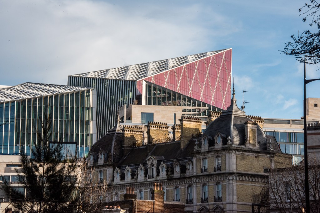 Nova South, a contemporary office building with distinctive pink and gray angular facade, rises behind a Victorian stone building in Victoria, London.