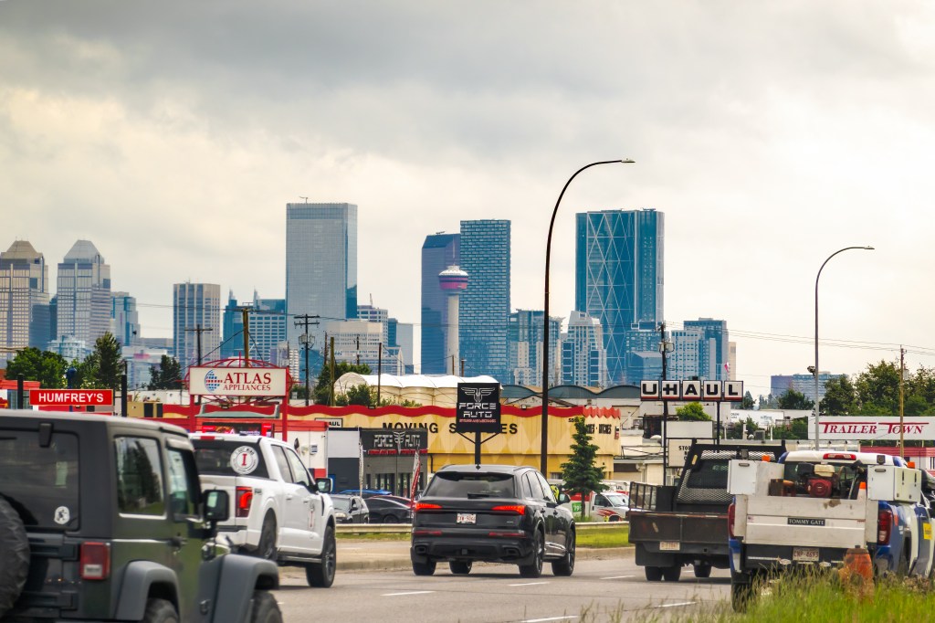 Vehicles driving through a commercial district with the downtown skyline and Calgary Tower behind