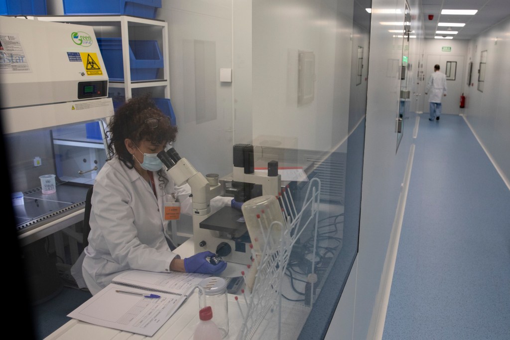 A photo of a white, female clinician wearing a mask and looking into microscope in a laboratory.
