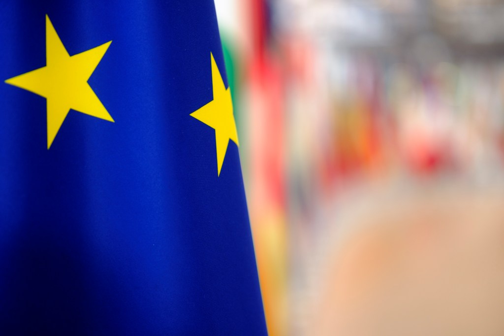 A photo of a detail of the flag of the European Union (Blue background with two gold stars) on the left side on a blurred background.