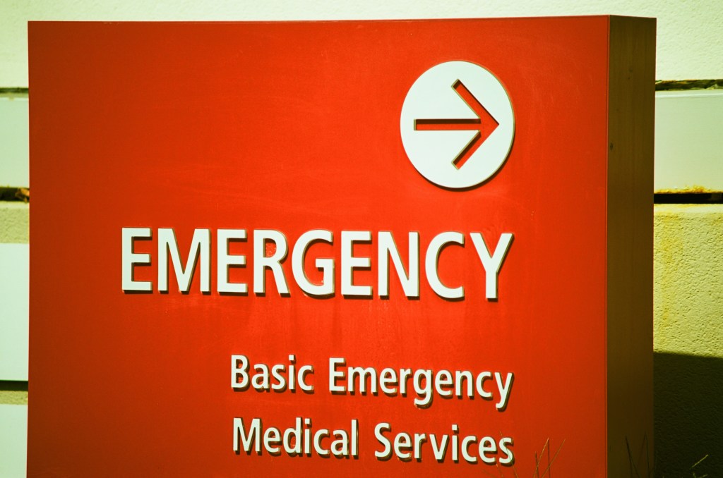 A photo of an emergency department sign at a hospital in white letters on a red field. There is an arrow above the word "Emergency."