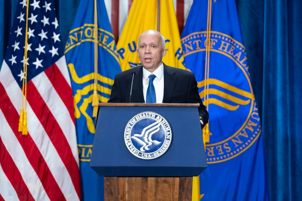 A photo of the Secretary of Labor speaking at a podium with the seal of the Department of Health and Human Services on it, flanked by numerous HHS flags (yellow and blue)