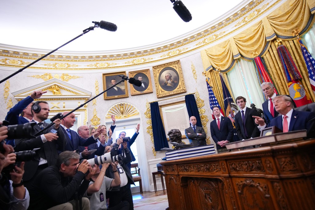 A photo of President Trump in seated behind the Resolute Desk in the Oval Awful surrounded by administration officials while speaking to a large number of reporters.