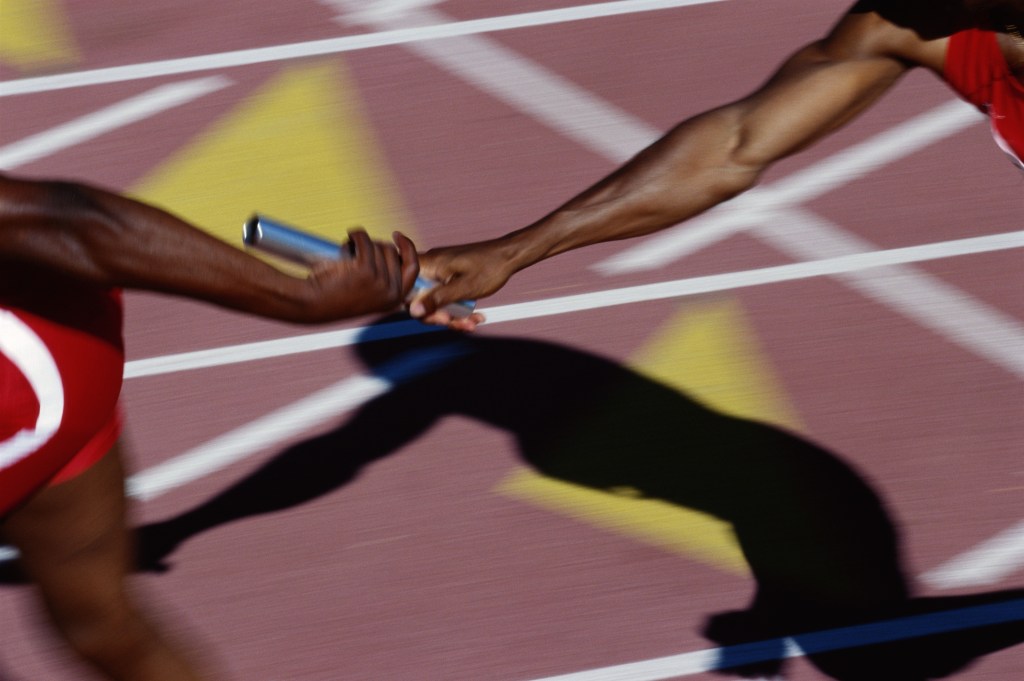 A photo of the arms of two brown-skinned runners passing a baton on a brick-red running track.