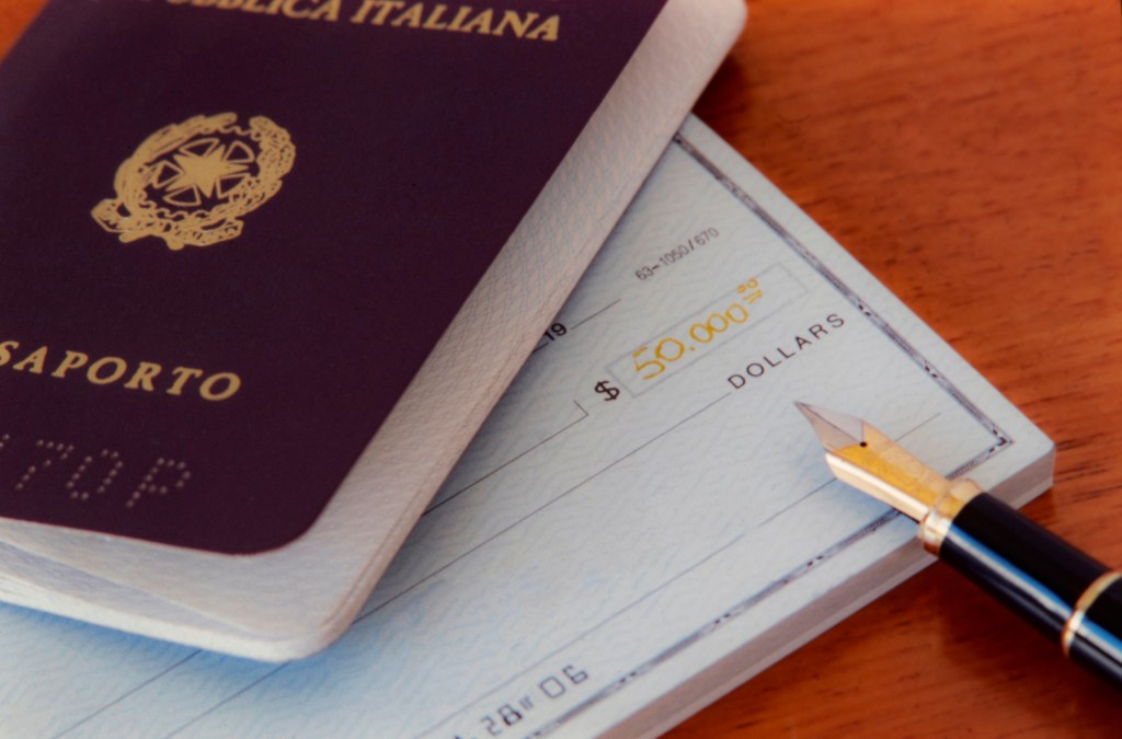 A close up photo of an Italian passport, a checkbook and a fountain pen on a wooden desk.