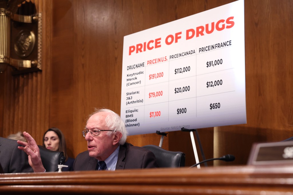 A photo of Senator Bernie Sanders seating below a poster titled "Price of Drugs" during a congressional meeting.