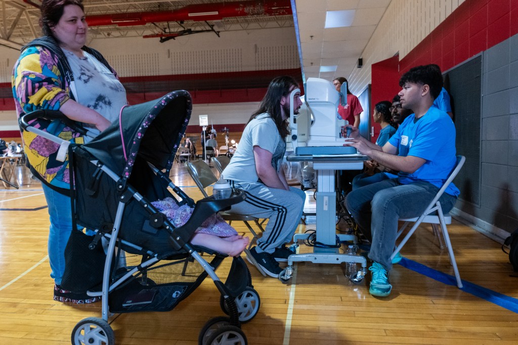 A photo of a medical clinic set up in a school gym with a woman and a baby stroller in the foreground and a man getting his eyes checked in the background.