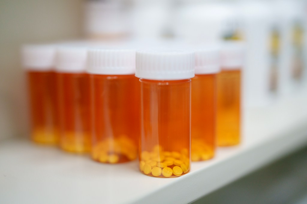 A photo of a dozen orange prescription drug bottes with white lids and no labels on a while shelf.