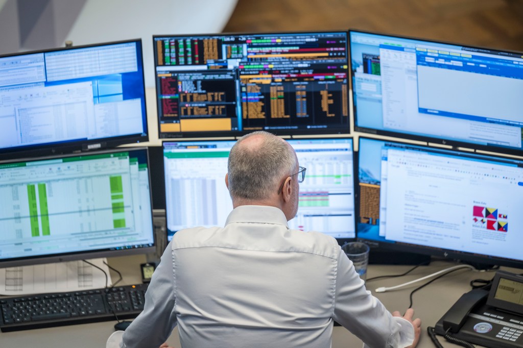 A photo of a back view of a seated white male with balding gray hair in front of six computer monitors displaying financial data.