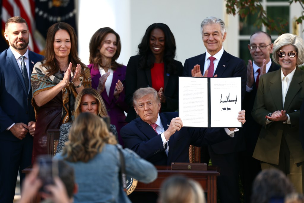 A photo of President Donald Trump holding up an executive order he just signed with a crowd of officials standing behind him, including CMS administrator Mehmet Oz and White House Chief of Staff Susie Wiles.