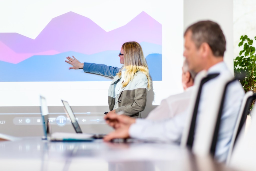 A female executive points to explain a graph in the background while other employees listen in the foreground at a table working on laptops.
